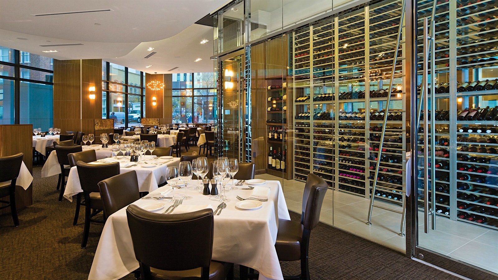 The wood-lined, modern-design dining room at Grace, with a wine room full of bottles, behind a glass panel